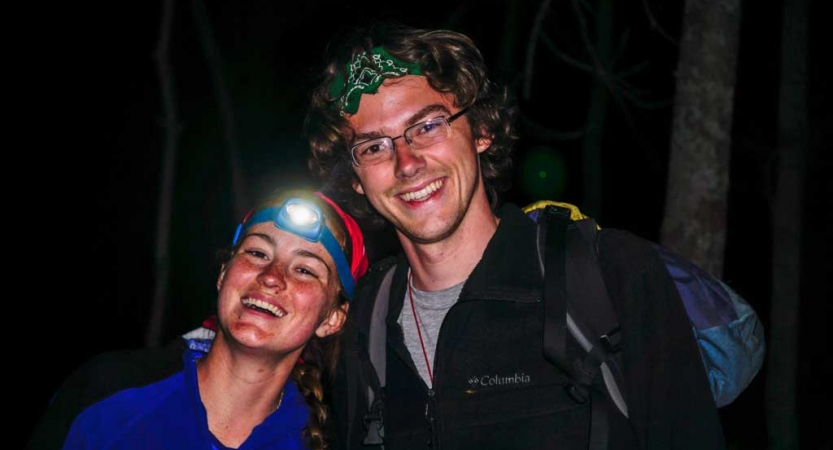 Two people smile for a photo at night. One of them is wearing an illuminated headlamp. 